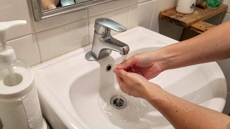 Hands being washed under running water at a small sink with a soap dispenser and a paper towel holder beside it.