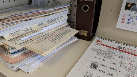 A stack of dated printed notices and a closed ring binder on a shelf, a wall calendar open beside them.