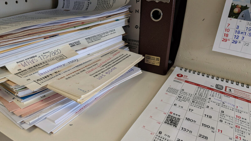A stack of dated printed notices and a closed ring binder on a shelf, a wall calendar open beside them.