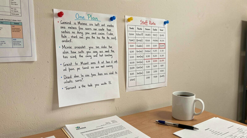 A handwritten one-page plan pinned to an office wall beside a staff rota, a mug and a pen on the desk below.