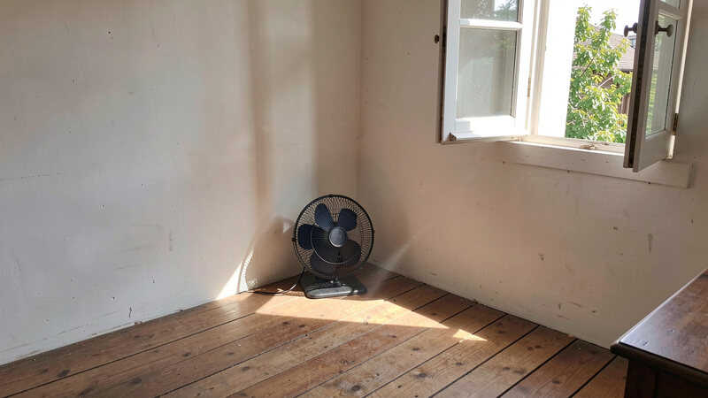 A room corner with an open window, a small fan placed low near the floor pointing towards the opening, sunlight on the boards.